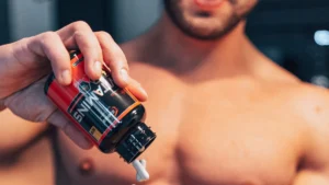 In a gym setting a man's hand holding a bottle of workout supplements and dispensing a pill. The man's torso is in the background, blurred and out of focus, emphasizing the action of the hand and the workout supplement bottle.